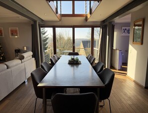 A modern dining area with a long table and chairs, large windows offering a view of the outdoors, a cozy seating area with a sofa, and a Christmas tree. - Cottage in the Heart of the Ardennes (Waimes)