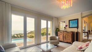 Ein geräumiges Wohnzimmer mit einem großen Glasschaufenster, das zu einem Pool führt, einer gemütlichen Couch, einer Holzhütte und Blick ins Freie.