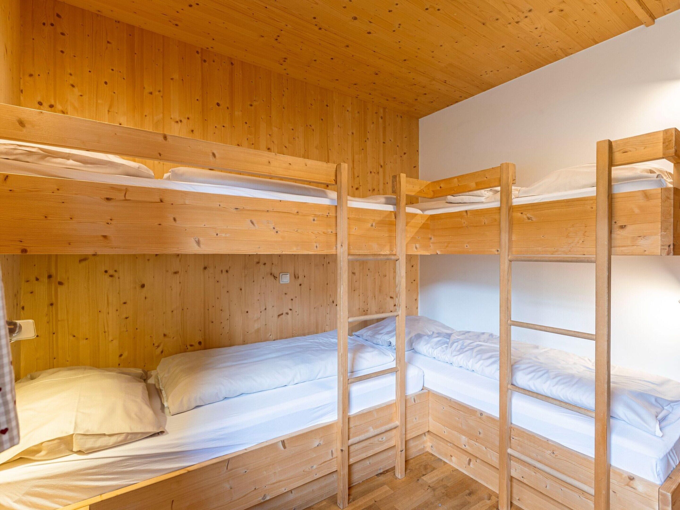 A bunk bed room with wooden walls and floors, a white ceiling, and a small bedside table with a lamp.