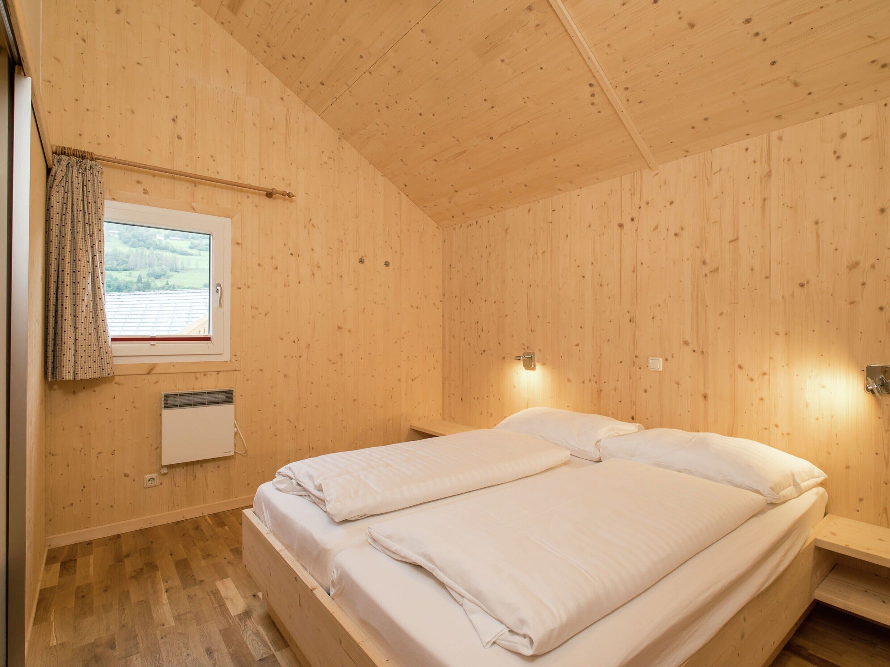 Una habitación con paneles de madera, una cama, un ventanal con cortinas, un calentador de pared y un estante pequeño.
