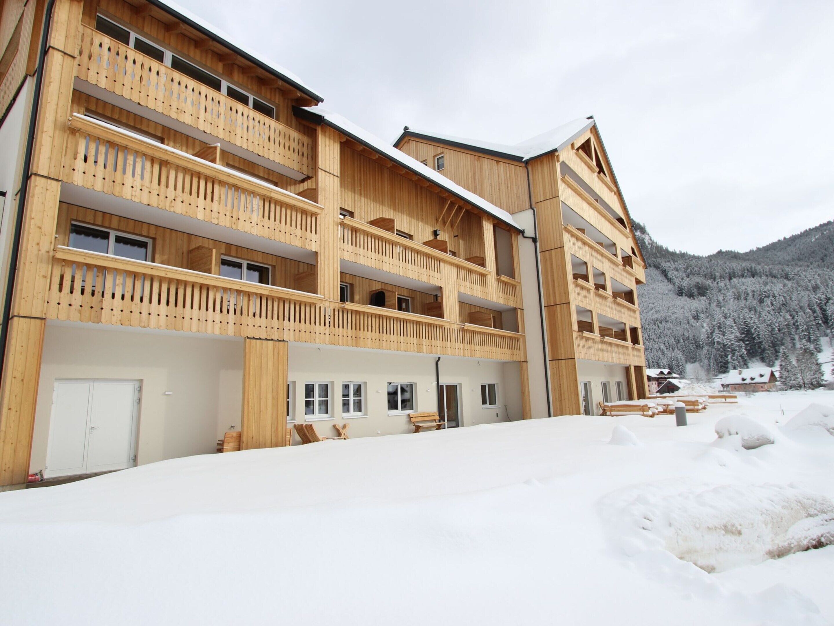 A wooden multi-story building with balconies, a flat roof, and a snowy landscape in the background.