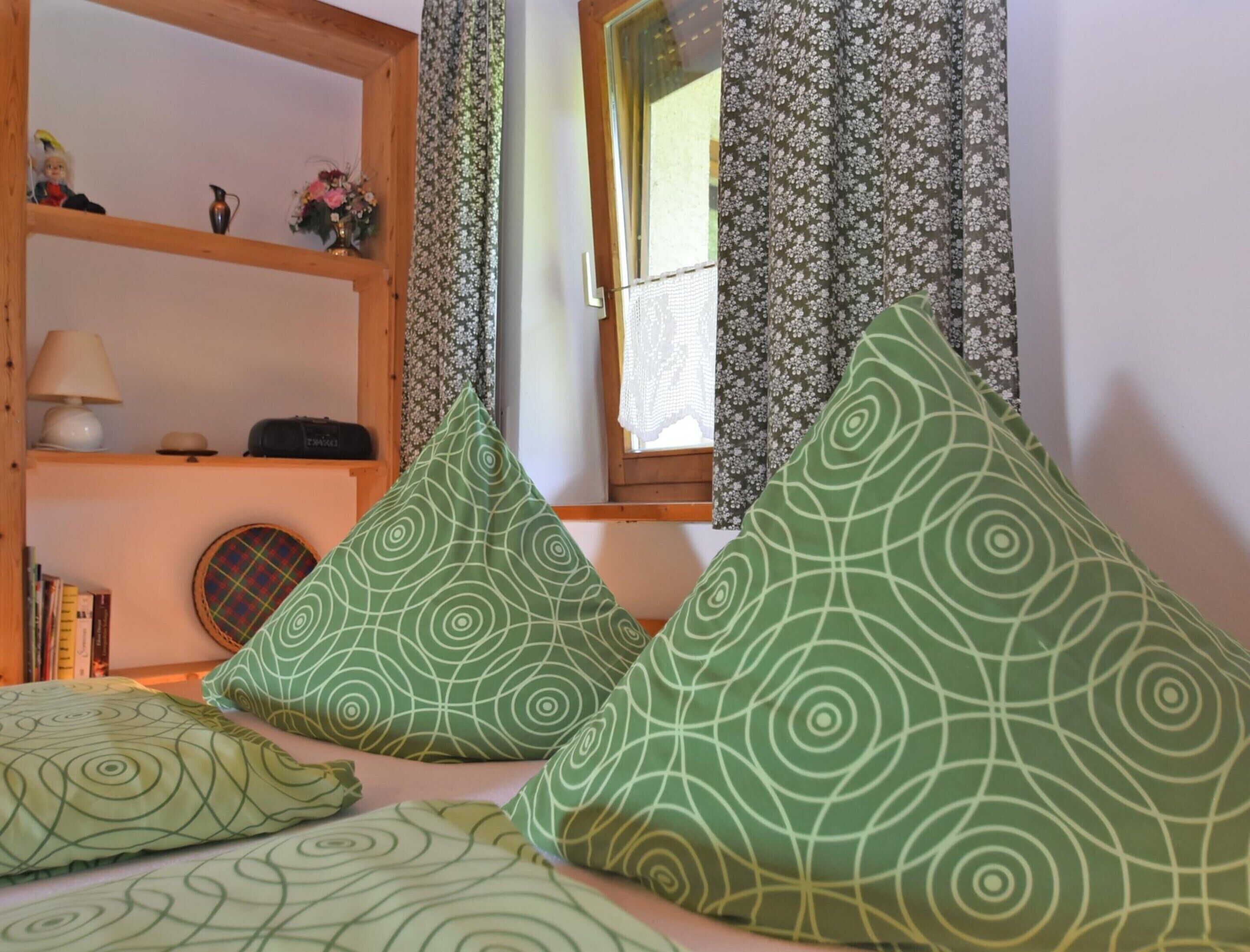 A bedroom with a bed, pillows, a shelf with books, a lamp, and a window with curtains.