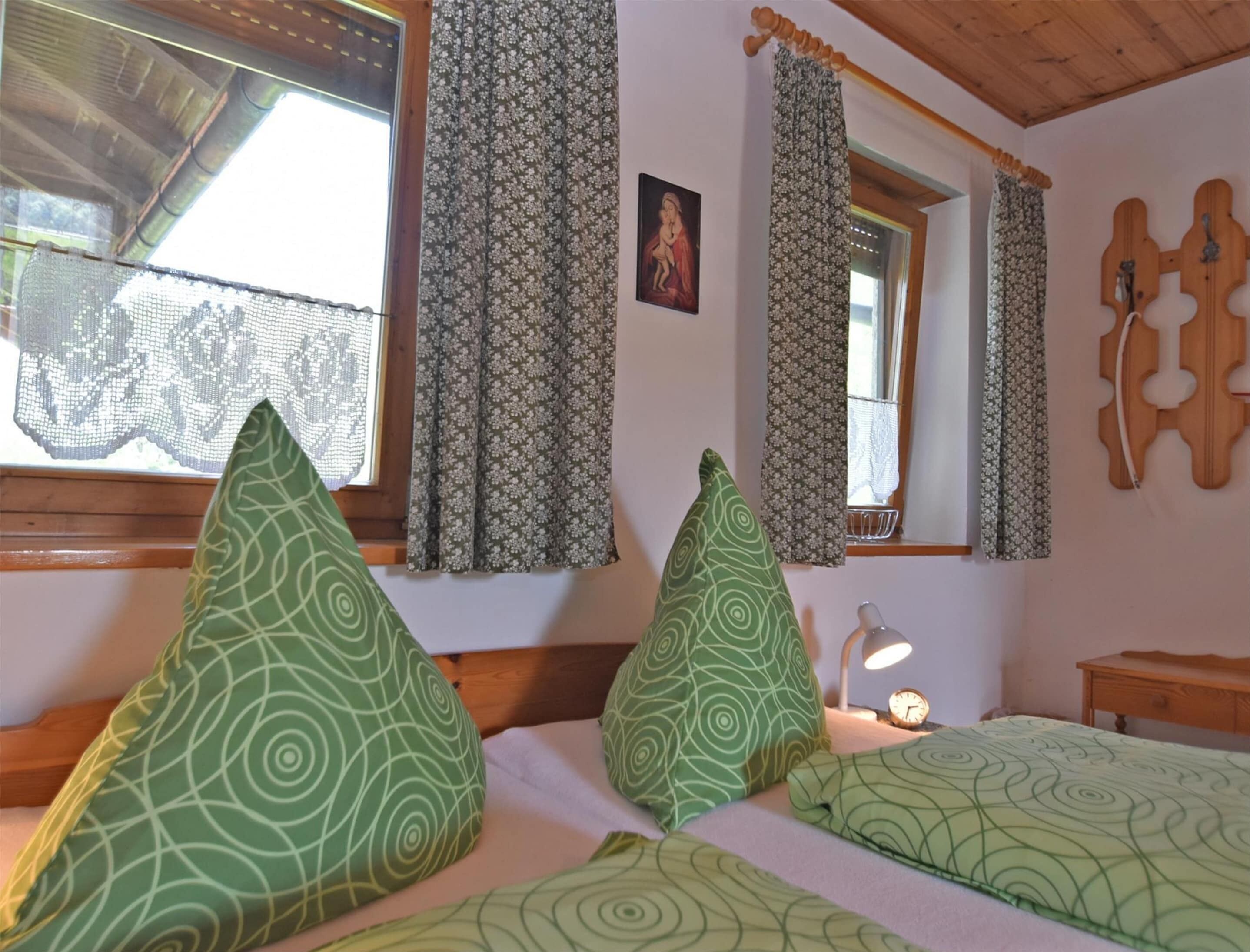 A room with two beds, each with green patterned pillows, a wooden headboard, lace curtains, a wooden ceiling, and a small bedside table with a lamp.