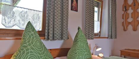 A room with two beds, each with green patterned pillows, a wooden headboard, lace curtains, a wooden ceiling, and a small bedside table with a lamp.