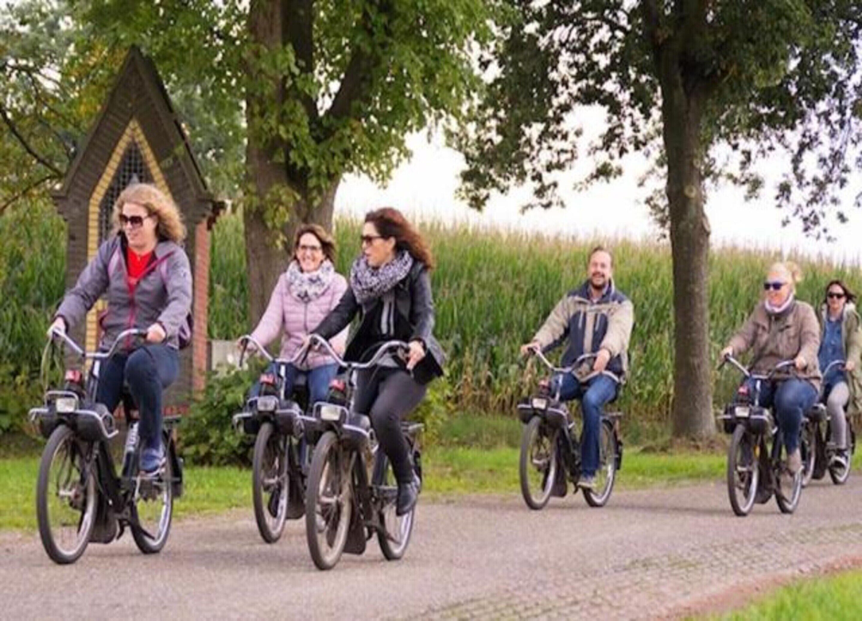 A group of people riding bicycles on a paved path with trees and a building in the background.