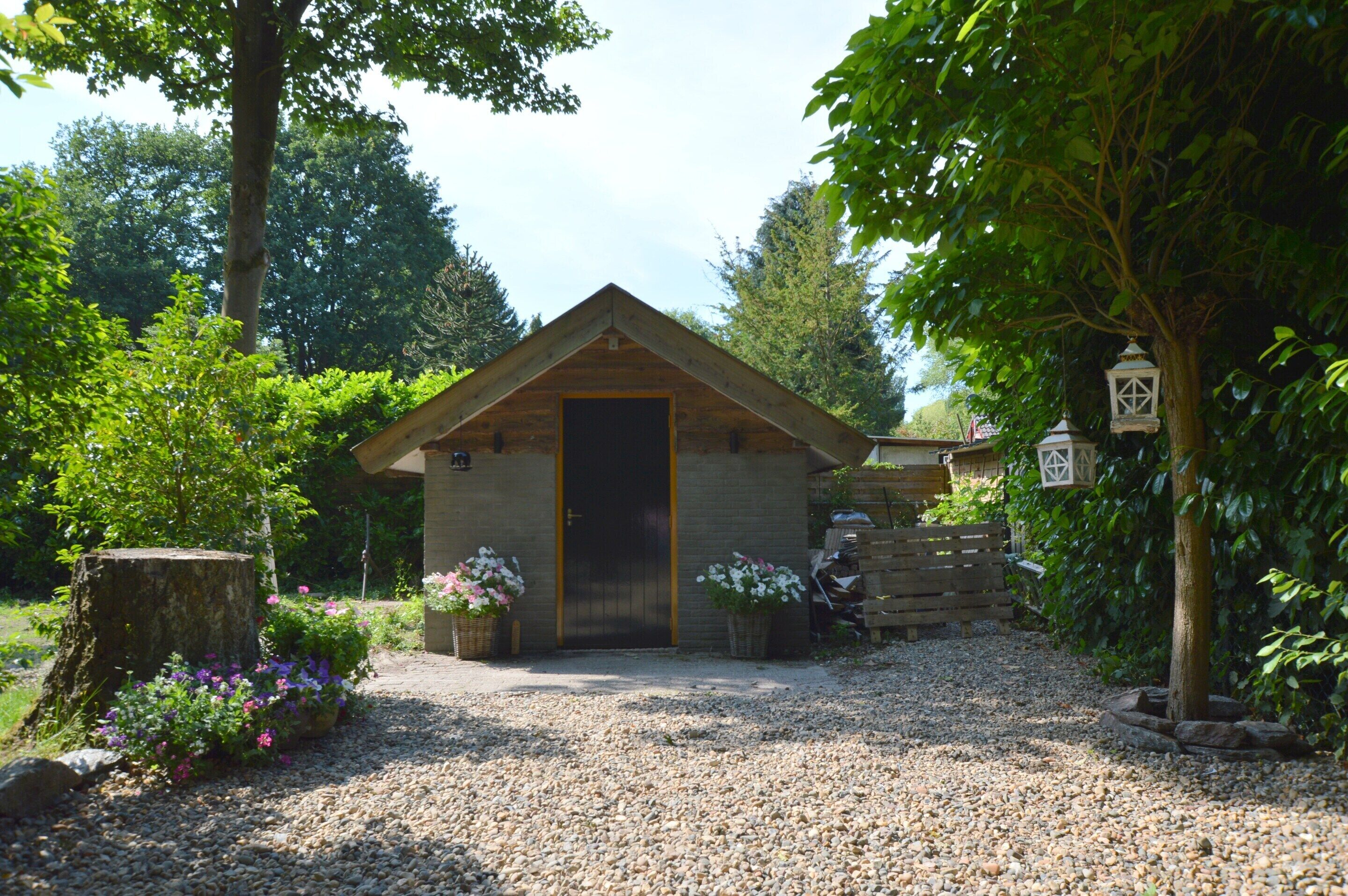 Une petite cabane en bois avec une porte, entourée de gravier, d’arbres et de parterres de fleurs.
