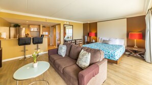A modern hotel room with a brown sofa, a glass coffee table, a bed with a blue patterned bedspread, and a red lamp.