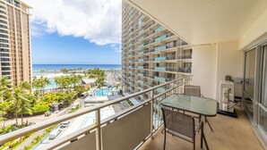 A balcony with a glass table and chair, overlooking a resort area with a pool, beach, and tall buildings.
