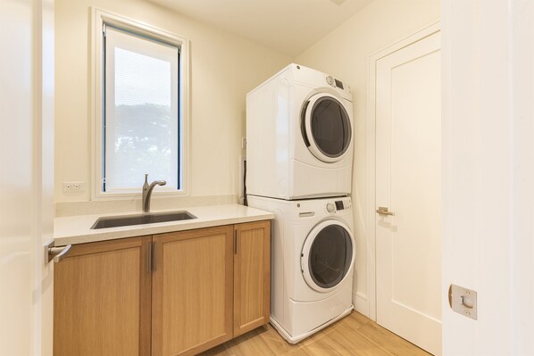 A laundry room with two stacked washing machines, a sink, wooden cabinets, and a window. - KLVR Presents Honua Kai Resort Luana Villas 11B&D (Lahaina)