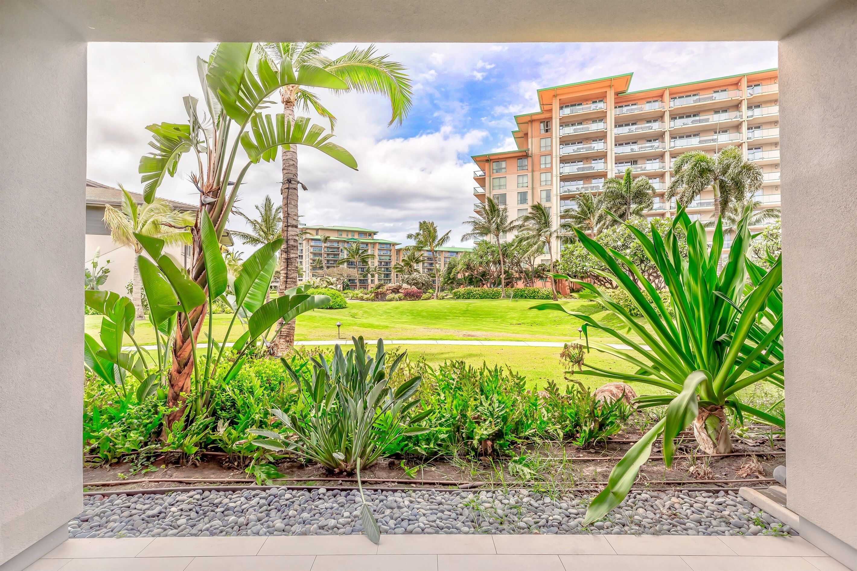 A hotel courtyard with lush greenery, a well-maintained lawn, and a view of a multi-story building in the background.