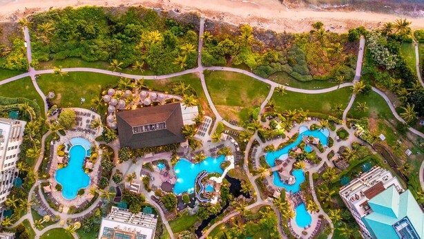 Luftaufnahme eines Resorts mit mehreren Schwimmbecken, üppiger Vegetation und einem Strand.