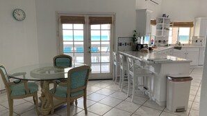 Una cucina con un tavolo da pranzo in vetro, sedie bianche e vista sull'oceano attraverso ampie porte.