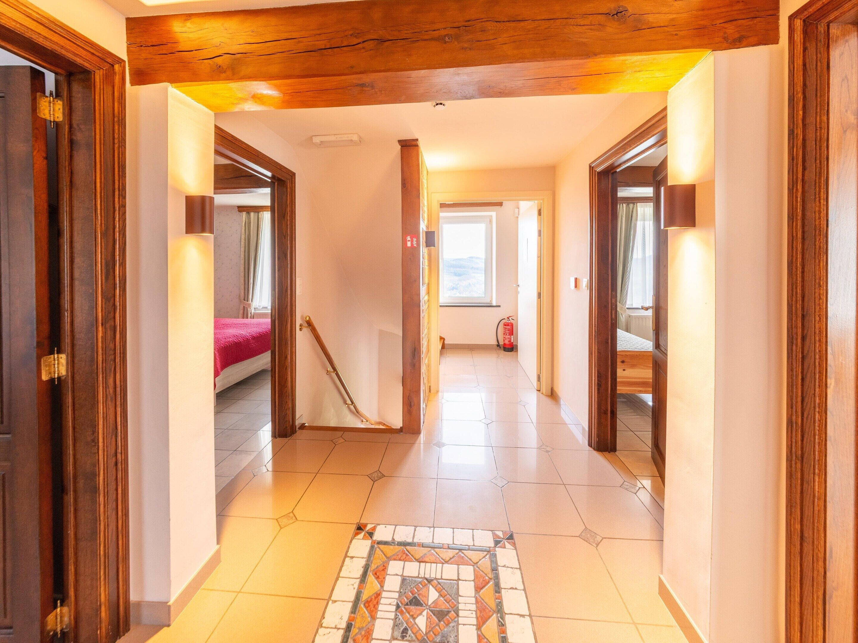 A hallway with wooden doors, a tiled floor, and a bed with a red blanket in the background.