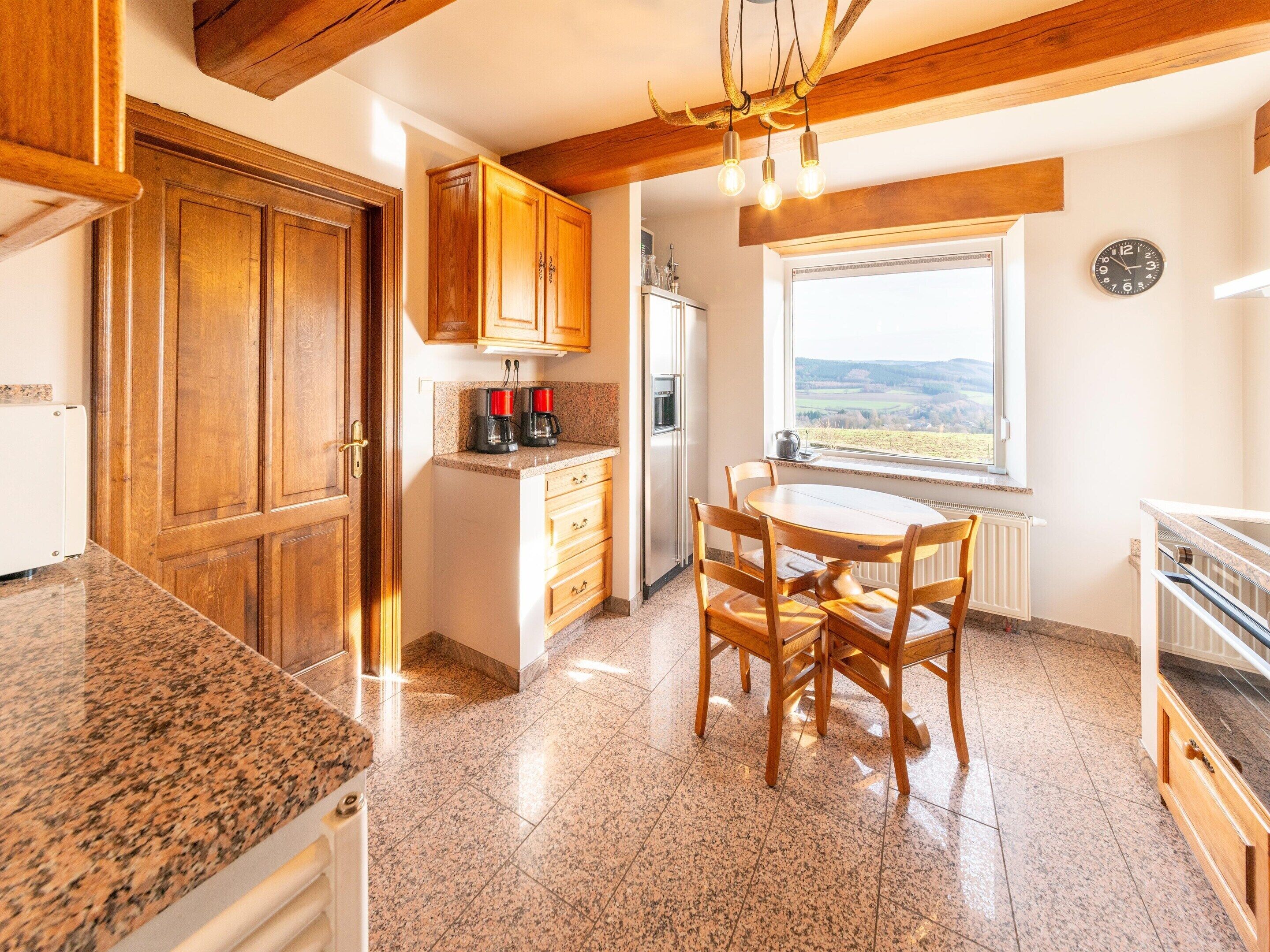 Une cuisine dotée de plans de travail en granit, de meubles en bois, d’une table à manger avec des chaises, d’un four encastré et d’une fenêtre offrant une vue sur la campagne.