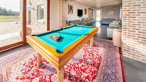 Een pooltafel in een ruimte met een rood behangtapijt, bakstenen muren en een groot raam met uitzicht op een terras.