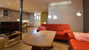 Un salon avec un téléviseur, un canapé et une table. Au fond, on aperçoit une cuisine avec un mur en briques et une fenêtre.