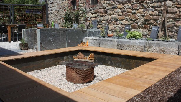 Un bâtiment en pierre avec un foyer au centre, entouré d’une terrasse en bois et d’un sol en gravier.