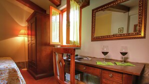 Een kamer met een houten bureau, twee stoelen, een bed met een bloemenprint beddegoed, een spiegel en twee glazen rode wijn op het bureau.