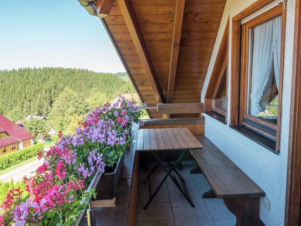 Un balcon en bois, agrémenté d’une table et de chaises, entouré de jardinières remplies de fleurs roses et violettes, et offrant une vue sur une zone boisée.