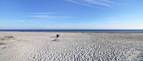 A wide sandy beach with tire tracks and a blue trash bin.