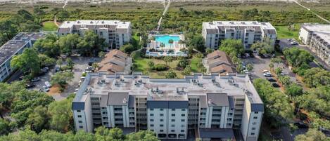 A hotel with a central pool, surrounded by lush greenery and a beach view.