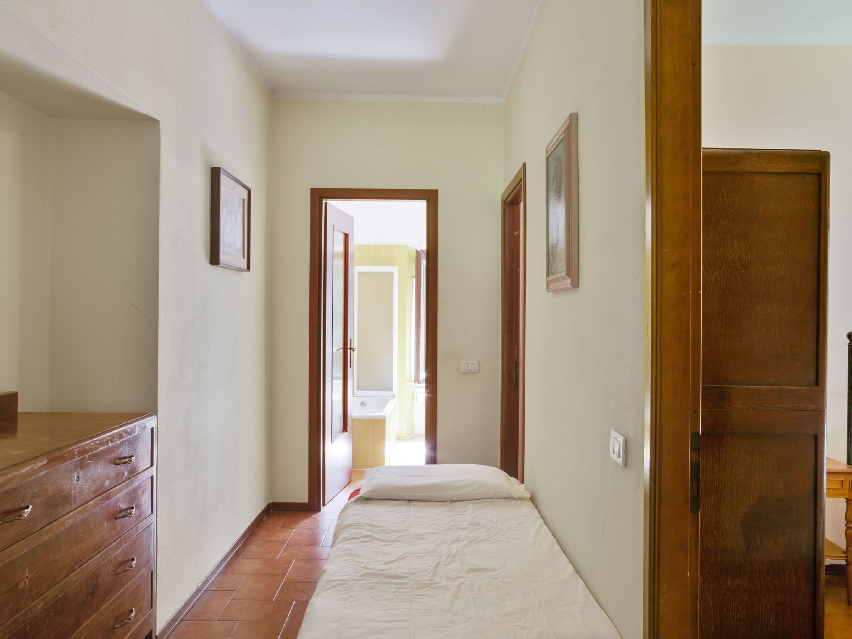 A narrow hallway with a bed at the end, wooden wardrobe, and a door leading to another room.