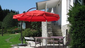 Un patio con una mesa, dos bancos, un paraguas rojo y una escalera de piedra que lleva a un edificio blanco.