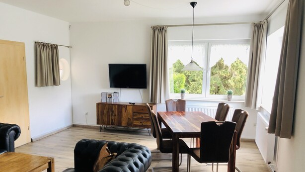 Una sala de estar moderna con un sofá de cuero negro, una mesa de comedor de madera y sillas. Un televisor de pantalla plana está montado en la pared, y hay ventanas con cortinas.