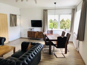 A modern living room with a black leather sofa, wooden dining table, and chairs. A flat-screen TV is mounted on the wall, and there are windows with curtains. - Cozy Holiday Home In Brilon near Ski Slopes (Brilon)
