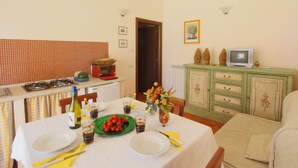 A dining area with a table set for two, a green plate with cherry tomatoes, a vase with flowers, and a television on a cabinet.