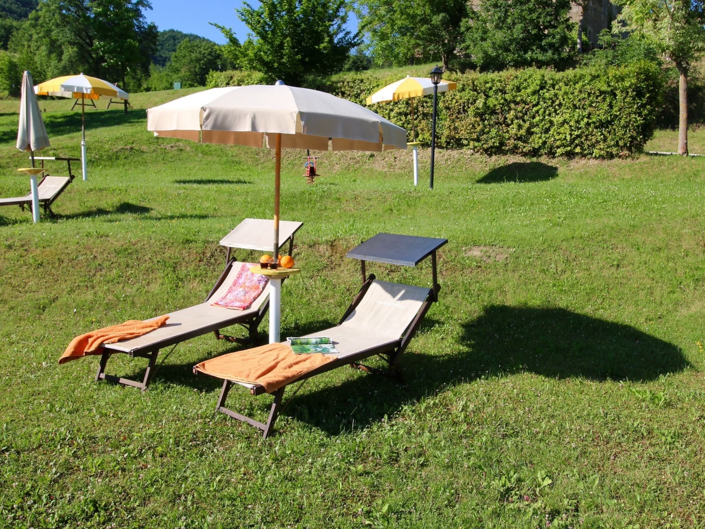 A grassy outdoor area with two sun loungers, one with a towel and orange cloth, and a table with a bottle and oranges. There are umbrellas and a lamp post in the background.