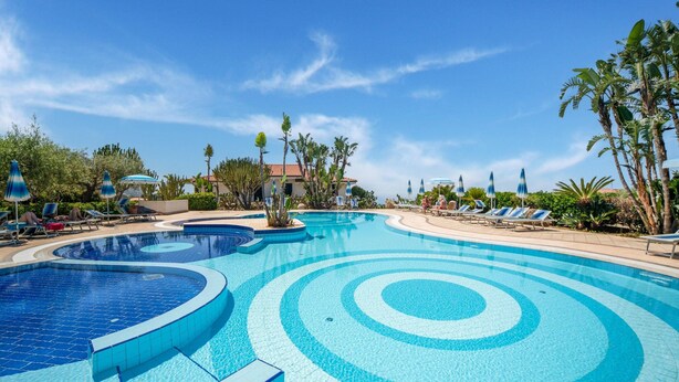 A large, multi-tiered swimming pool with clear blue water, surrounded by lounge chairs and umbrellas, and palm trees. - Belvilla by OYO Gattotrilo x 5 (Ricadi)