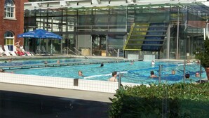 A swimming pool area with multiple lanes, a covered seating area with umbrellas, and a modern building with large windows. - Nature & Wellness in Sohl (Bad Elster)