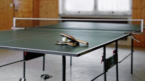 Une table de ping-pong avec une balle et une raquette au premier plan, le tout se détachant sur un mur en bois et une fenêtre en arrière-plan.