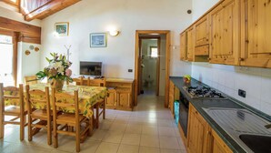 A kitchen with wooden cabinets, a dining table with chairs, a TV, and a bathroom visible through an open door. - Apartment in Antey-saint-andre Near Ski Lifts (Antey-Saint-Andre)