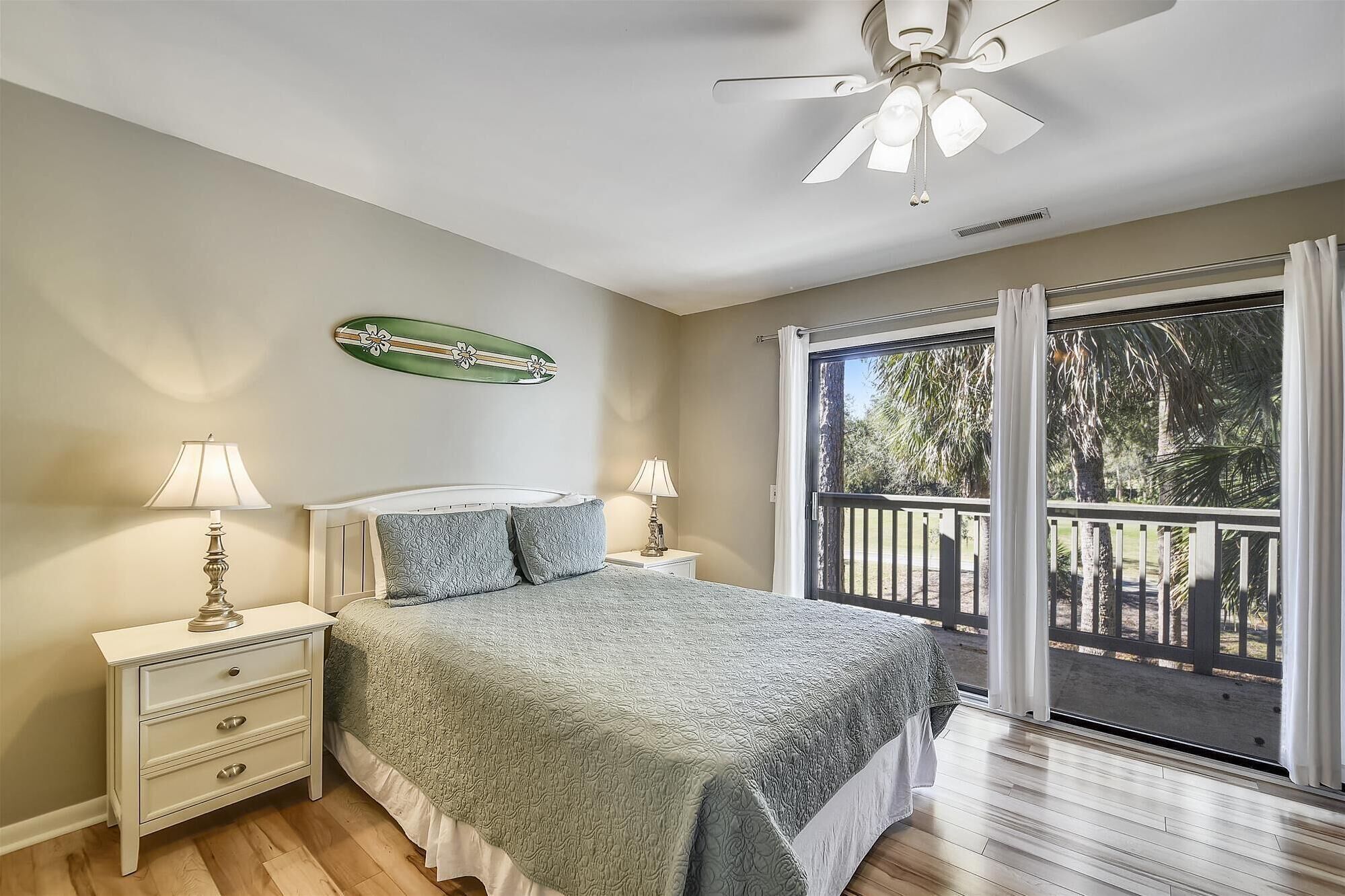 Um quarto com cama, duas luminárias de cabeceira, mesa de cabeceira, ventilador de teto, janela com vista para palmeiras e um objeto decorativo na parede.