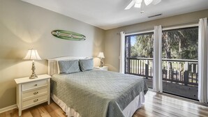 A bedroom with a bed, two bedside lamps, a nightstand, a ceiling fan, a window with a view of palm trees, and a decorative wall piece. - Sunset Rentals | Tennismaster 801 | Dog Friendly (Hilton Head Island)