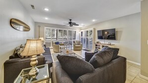 A spacious living room with a ceiling fan, a television mounted on the wall, and a large window with blinds. - Sunset Rentals | Tennismaster 801 | Dog Friendly (Hilton Head Island)