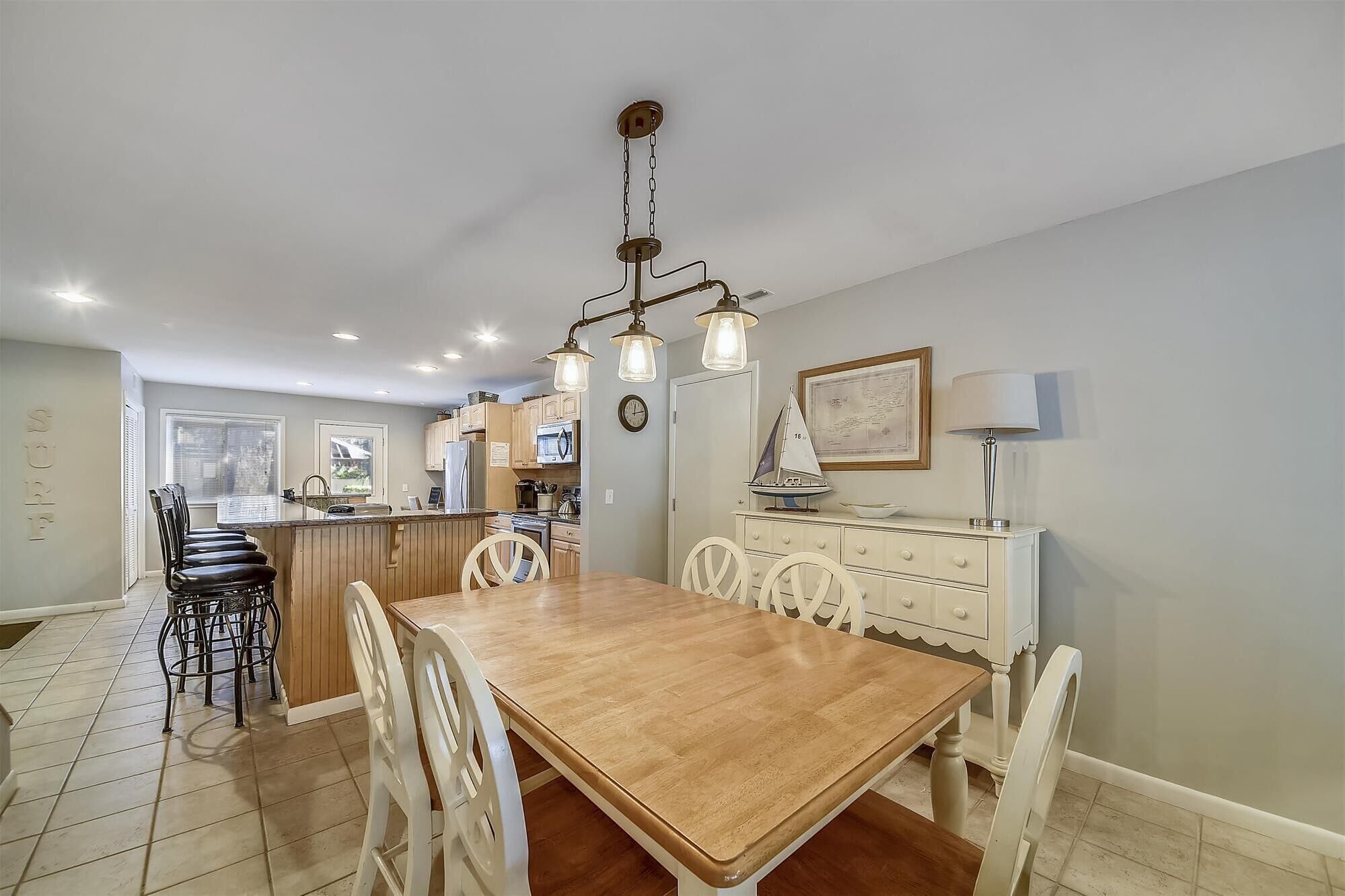 A dining area with a wooden table and white chairs, a kitchen with wooden cabinets, and a pendant light fixture above the table.