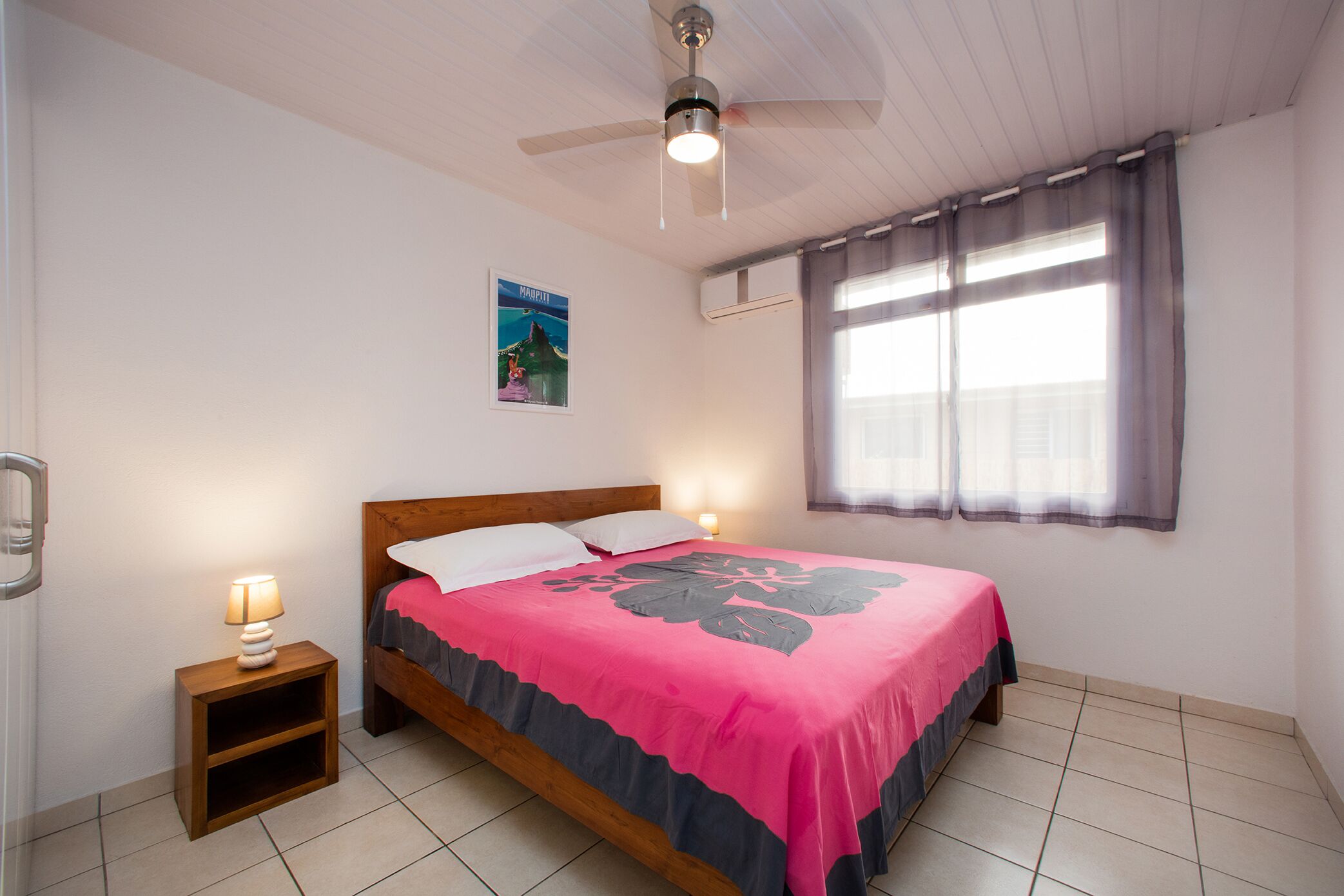 Ein Schlafzimmer mit Bett, Nachttisch, Deckenventilator, Fenster mit Vorhängen und einem Poster an der Wand.