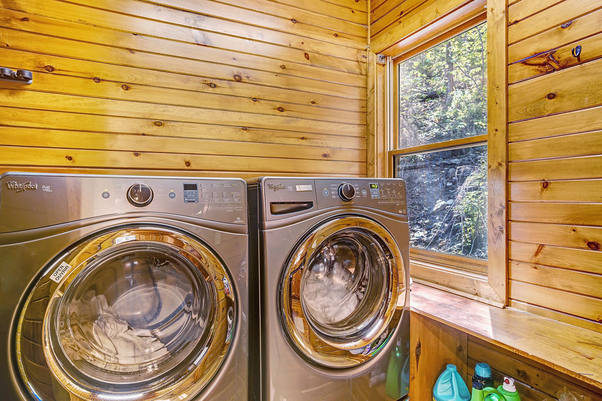 Two front-loading washing machines in a wooden cabin with a window overlooking a forest.