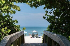 Ein Holzsteg führt zu einem Strand mit Menschen und Sonnenschirmen.