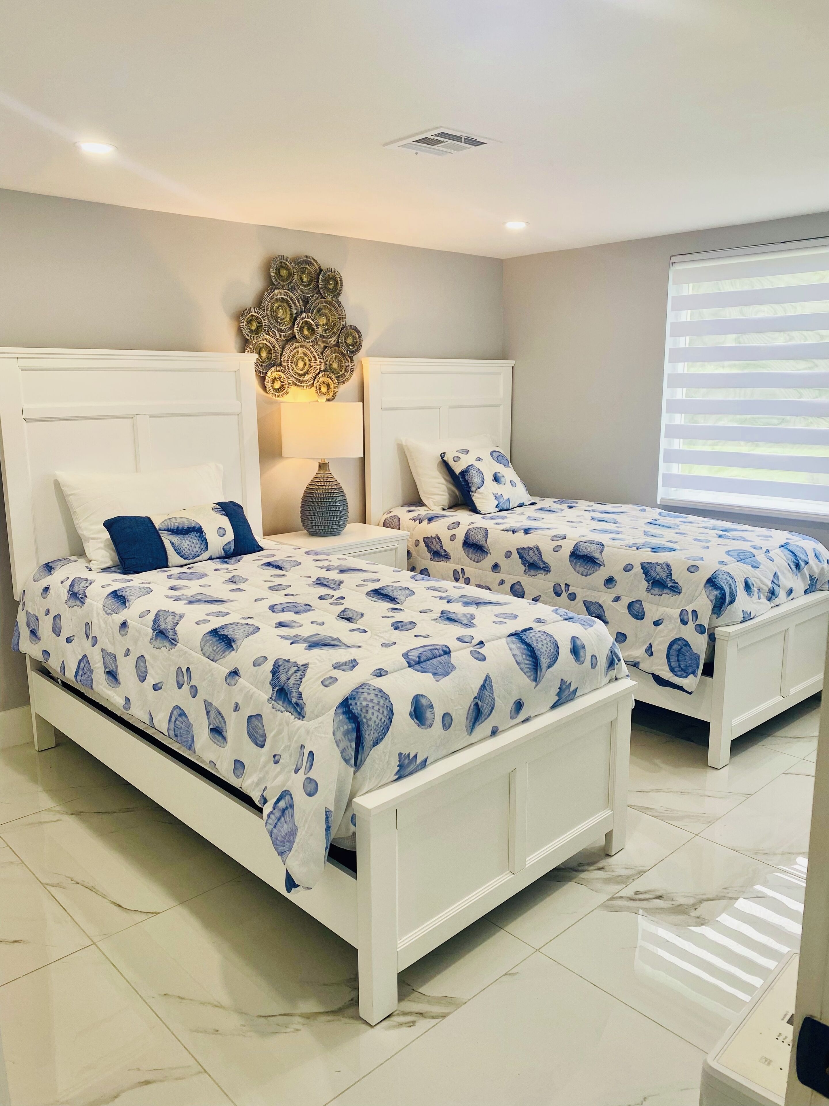 A bedroom with two white beds, each with a blue and white patterned bedspread. There is a decorative wall piece above the beds and a window with blinds on the right.