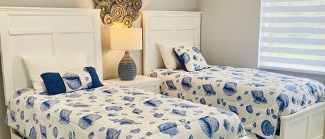 A bedroom with two white beds, each with a blue and white patterned bedspread. There is a decorative wall piece above the beds and a window with blinds on the right.