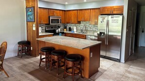 A modern kitchen with wooden cabinets, a stainless steel refrigerator, a granite countertop, and a bar stool area.