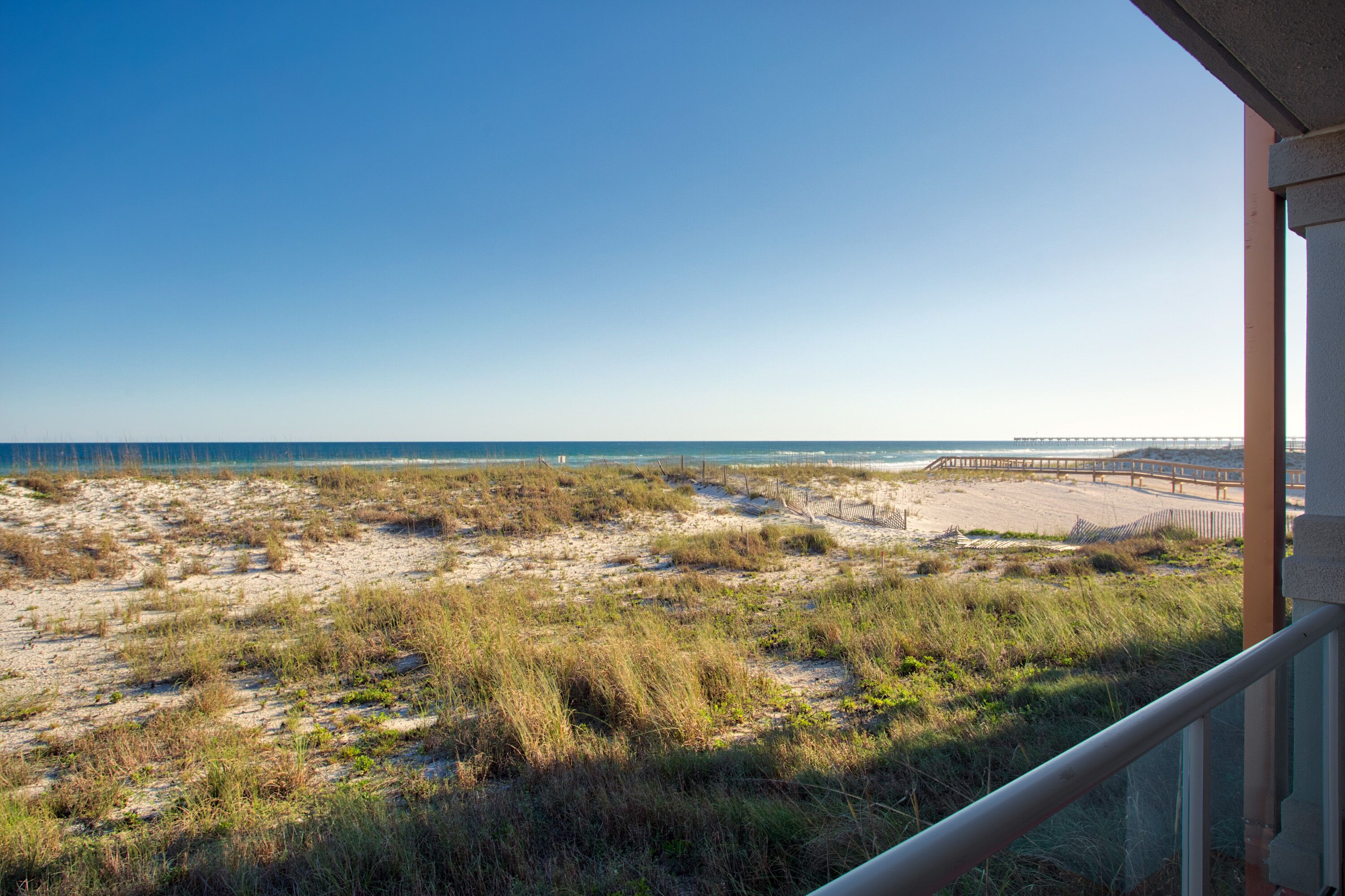 Strandudsigt med en træbro, der strækker sig ud i havet, sandklitter og en bygning med balkon.