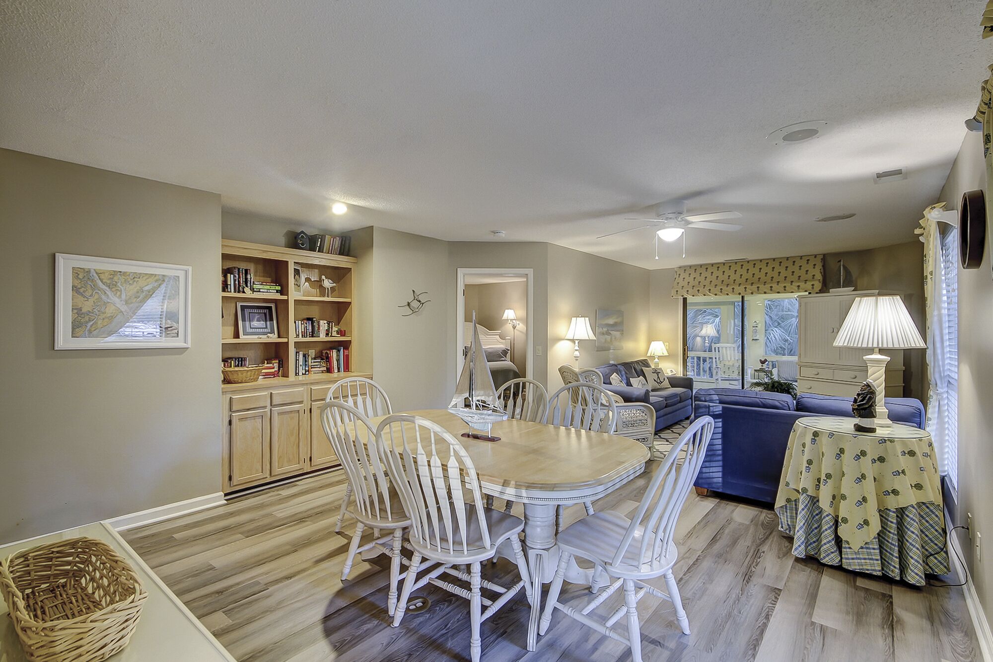 A dining area with a wooden table and white chairs, a bookshelf, and a living room with a sofa and lamps.