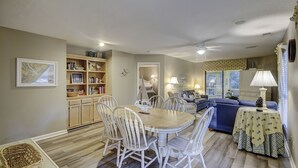 A dining area with a wooden table and white chairs, a bookshelf, and a living room with a sofa and lamps. - Sunset Rentals | Kingston Dunes 91 | Dog Friendly (Hilton Head Island)
