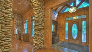 A log cabin interior with stone walls, wooden floors, and a large wooden door with stained glass. - Black Bear River Lodge | Pet Friendly | Hot Tub | Creek Access (Epworth)