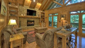 A log cabin interior with a stone fireplace, wooden furniture, and large windows. - Black Bear River Lodge | Pet Friendly | Hot Tub | Creek Access (Epworth)